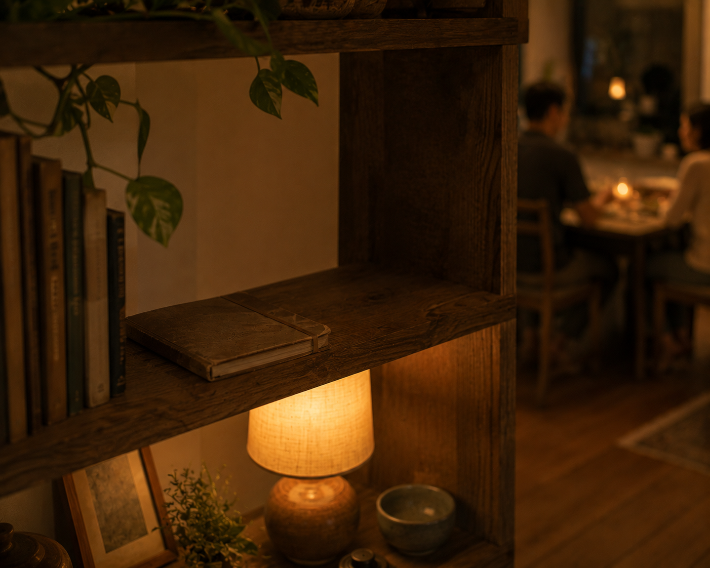 A single well-worn notebook resting on a shelf in warm evening light, two people sharing a relaxed meal blurred in the background.